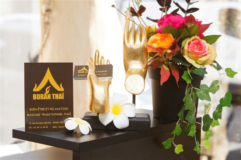 Présentation de l'accueil de BuranThaï, un centre de massage Thaï traditionnel réputé à Orléans. Des mains dorées dont l'une tient des cartes de visite.